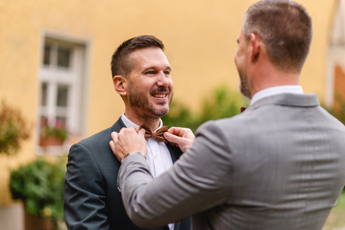 Zwei Männer in Anzügen stehen draußen. Einer rückt die Fliege des anderen zurecht. Sie lächeln vor einer gelben Wand mit einem Fenster.