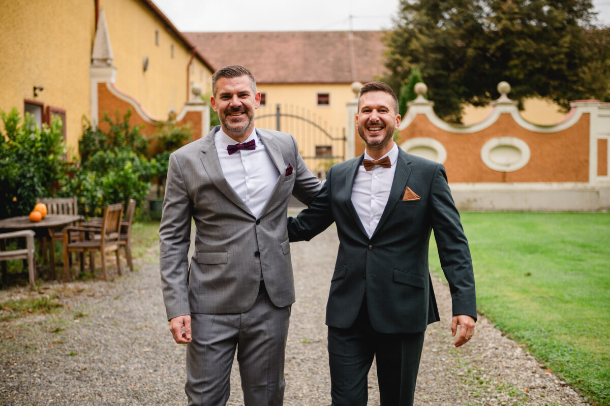 Zwei Männer in Anzügen gehen lächelnd auf einem Kiesweg in der Nähe eines gelben Gebäudes mit Garten. Einer trägt einen grauen Anzug, der andere einen dunklen Anzug, beide mit Fliege.