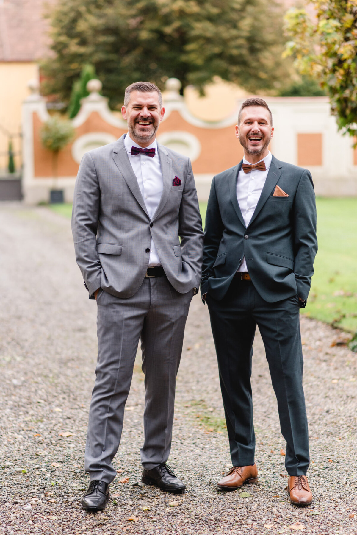 Zwei Männer in Anzügen stehen draußen auf einem Kiesweg und lächeln. Einer trägt einen grauen Anzug mit einer burgunderfarbenen Fliege, der andere einen dunklen Anzug mit einer braunen Krawatte. Im Hintergrund sind Bäume und eine dekorative Wand zu sehen.