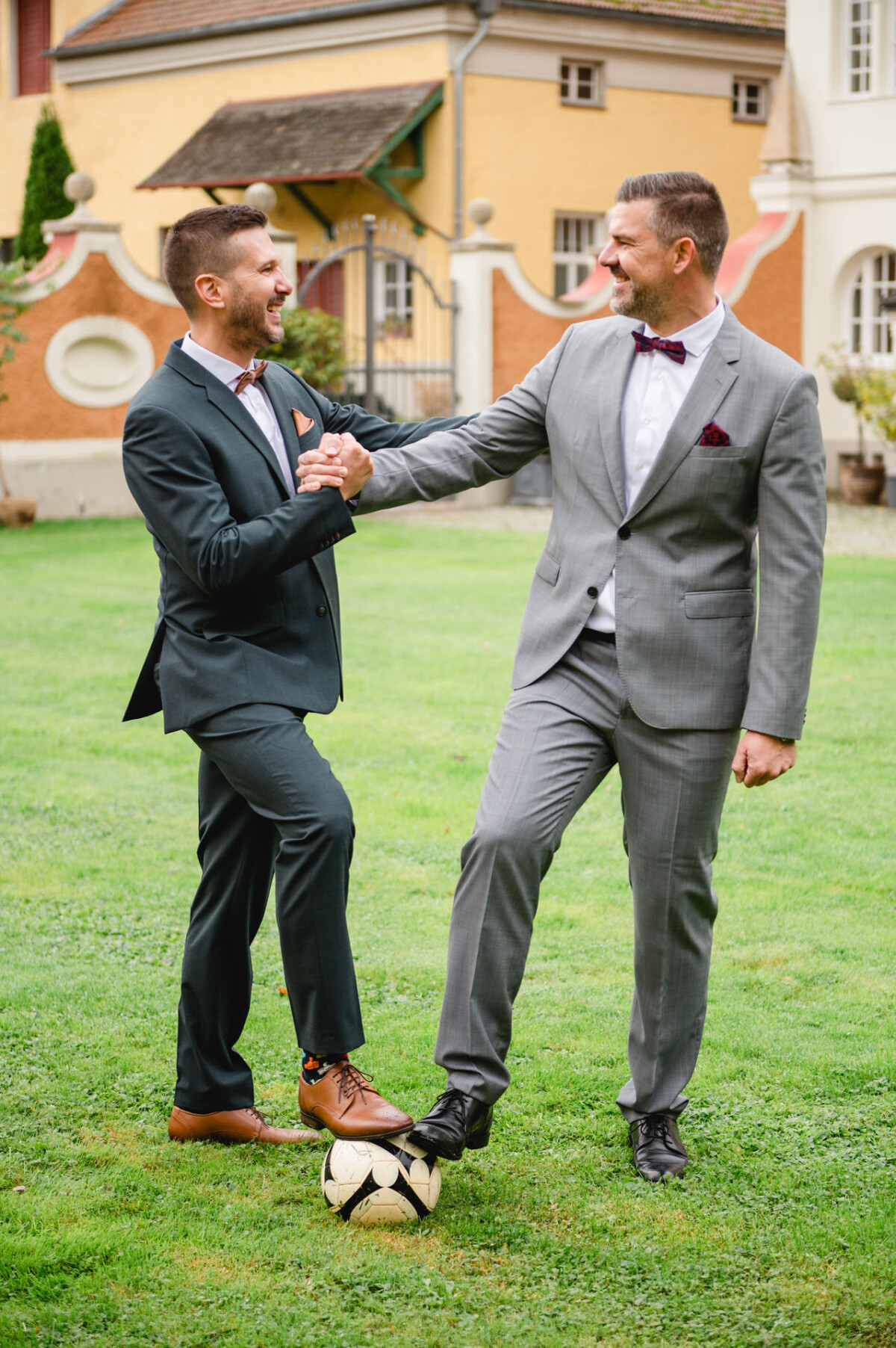 Zwei Männer in Anzügen geben sich die Hand, während sie auf einer Wiese stehen und zwischen ihnen ein Fußball liegt.