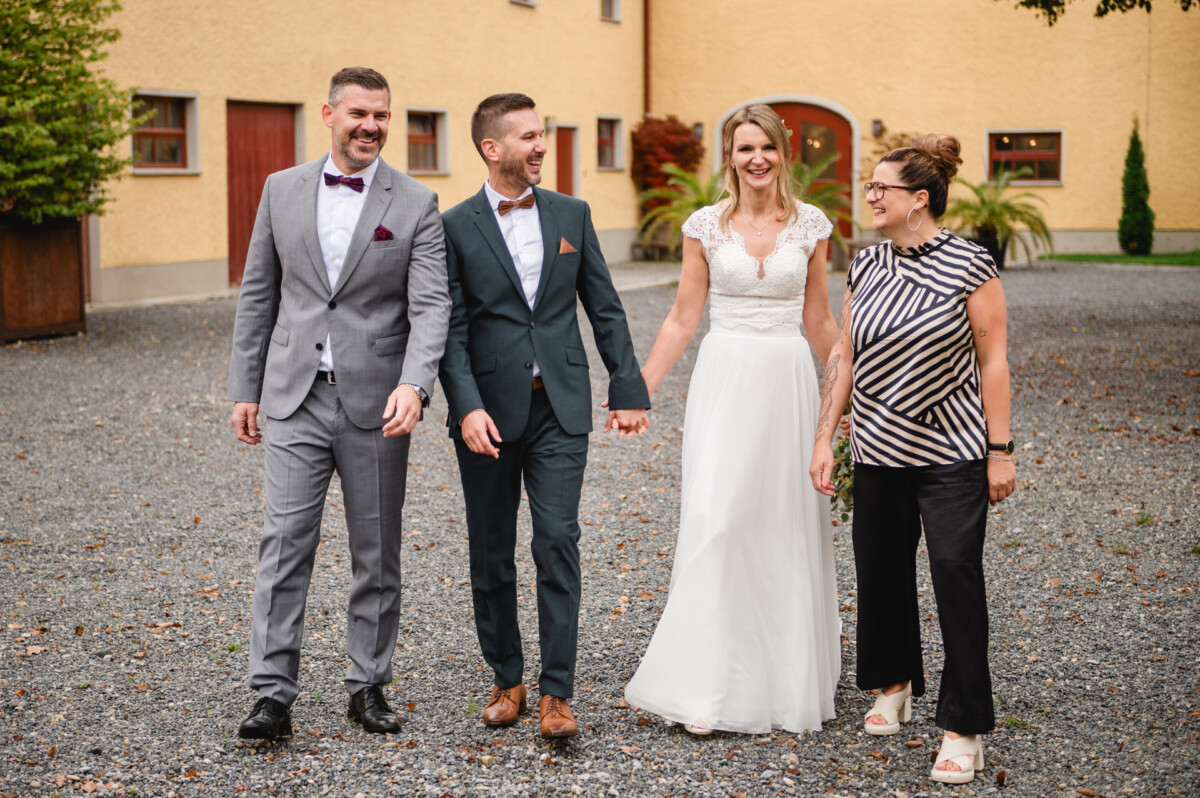 Zwei Männer in Anzügen, eine Frau im Brautkleid und eine weitere in Freizeitkleidung gehen gemeinsam auf einem Kiesweg.