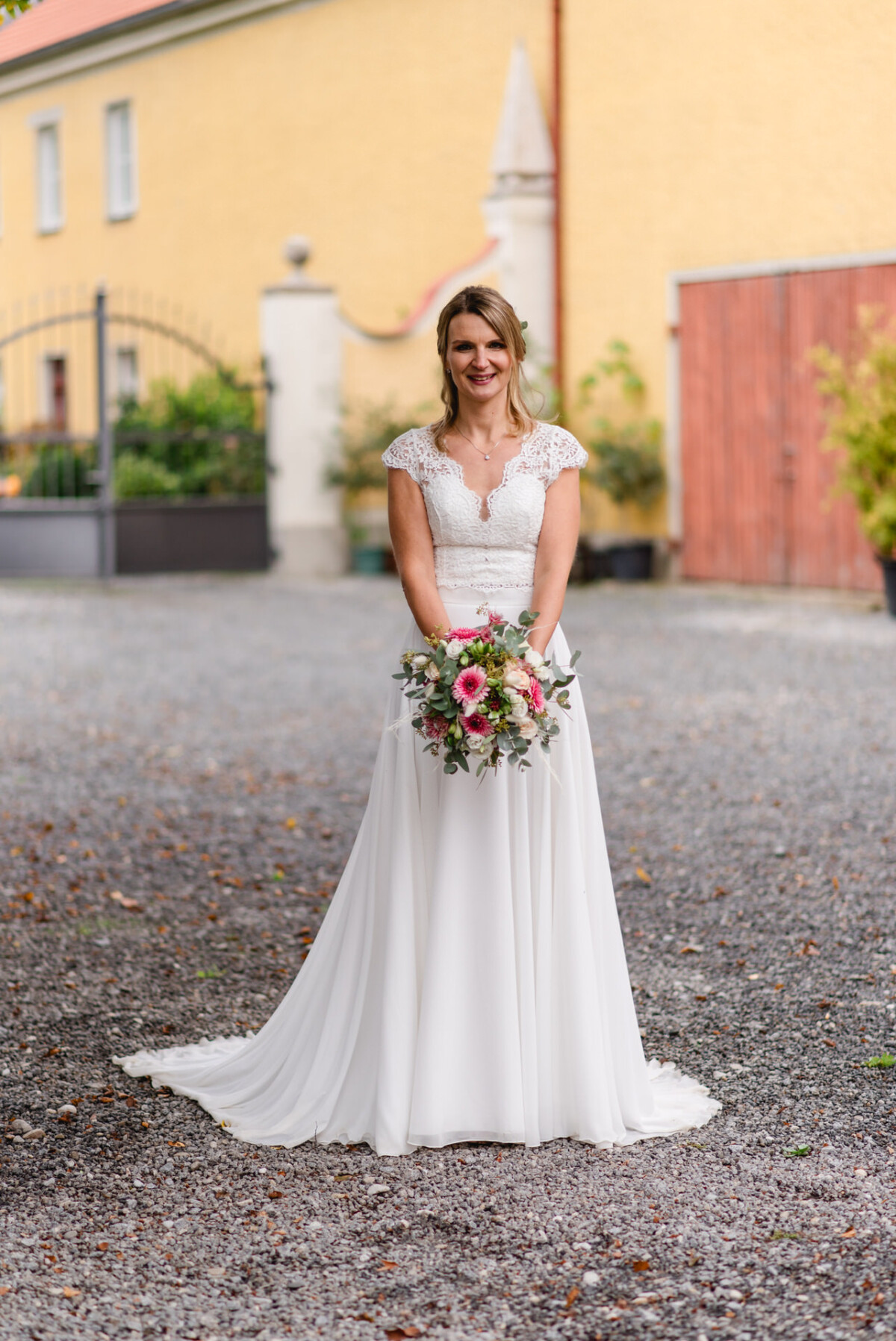 Eine Braut in einem weißen Hochzeitskleid hält einen Strauß rosa und weißer Blumen und steht auf einem Kiesweg mit Gebäuden im Hintergrund.