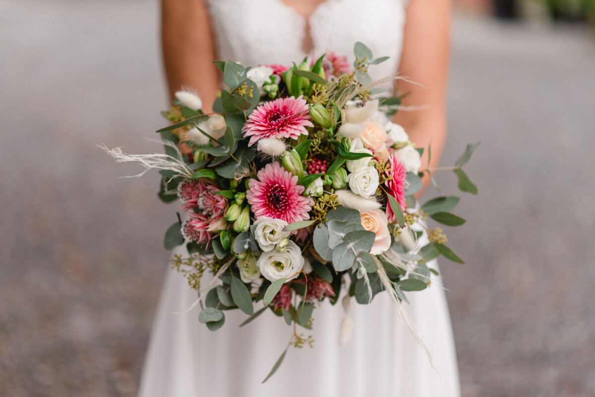 Eine Braut hält einen Strauß rosa und weißer Blumen mit Grün vor einem unscharfen Hintergrund im Freien.