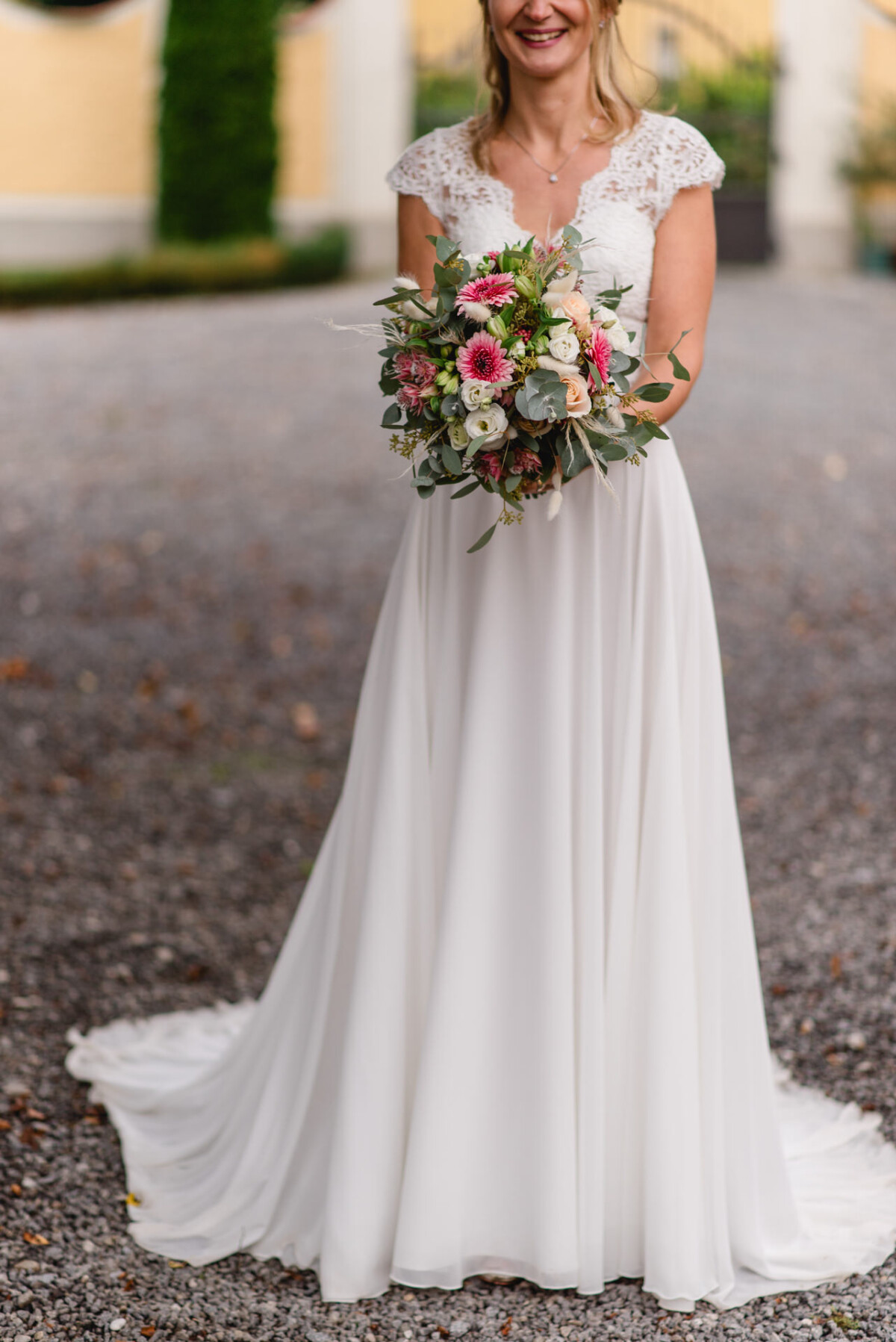 Eine Braut in einem weißen Kleid hält einen Strauß bunter Blumen und steht auf einem Kiesweg.