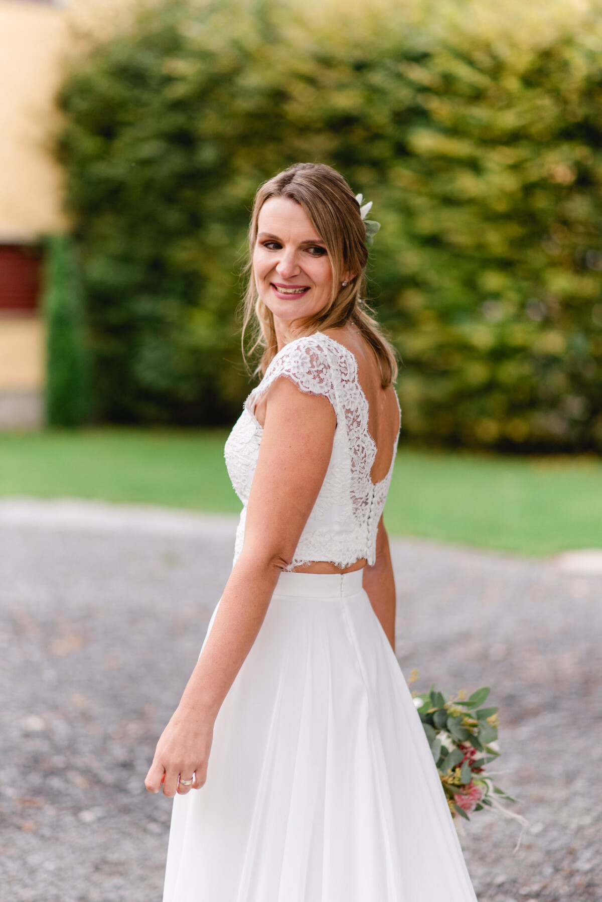 Eine Frau in einem weißen Spitzenbrautkleid steht im Freien, hält einen Blumenstrauß in der Hand und lächelt. Im Hintergrund ist Grün zu sehen.