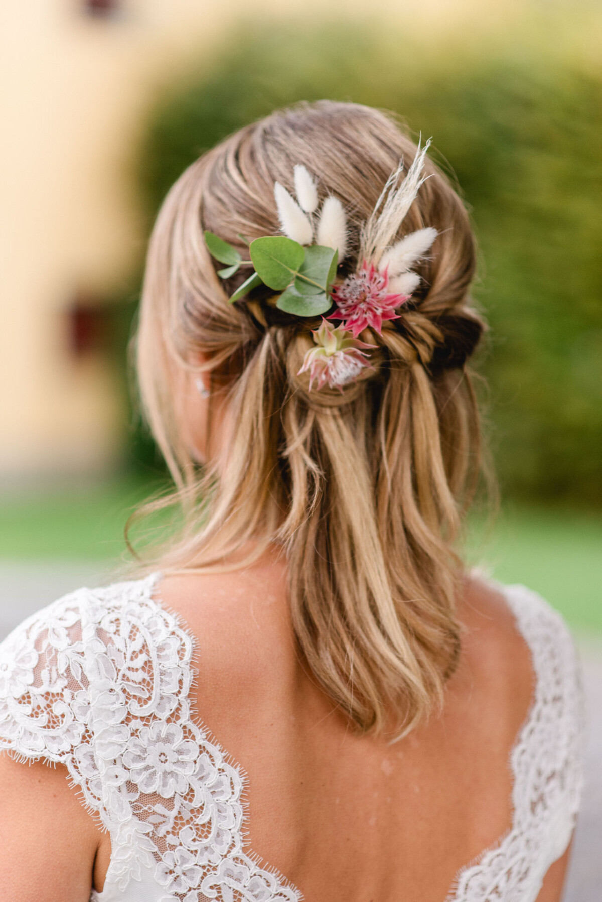 Von hinten ist eine Frau mit geflochtenem hellbraunem Haar, das mit Blumen und Grün geschmückt ist und ein weißes Spitzengewand trägt, zu sehen.