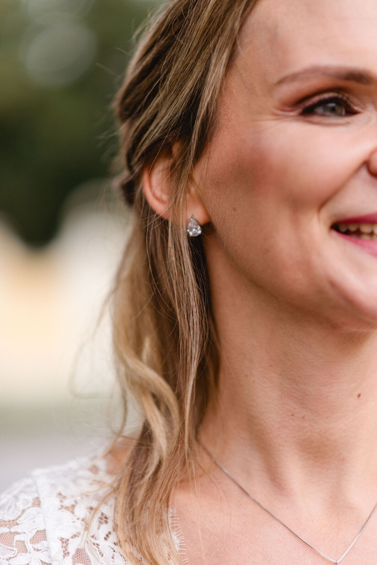 Nahaufnahme einer Frau mit langem, hellbraunem Haar, die ein weißes Spitzenkleid und eine zarte Halskette trägt und lächelt.