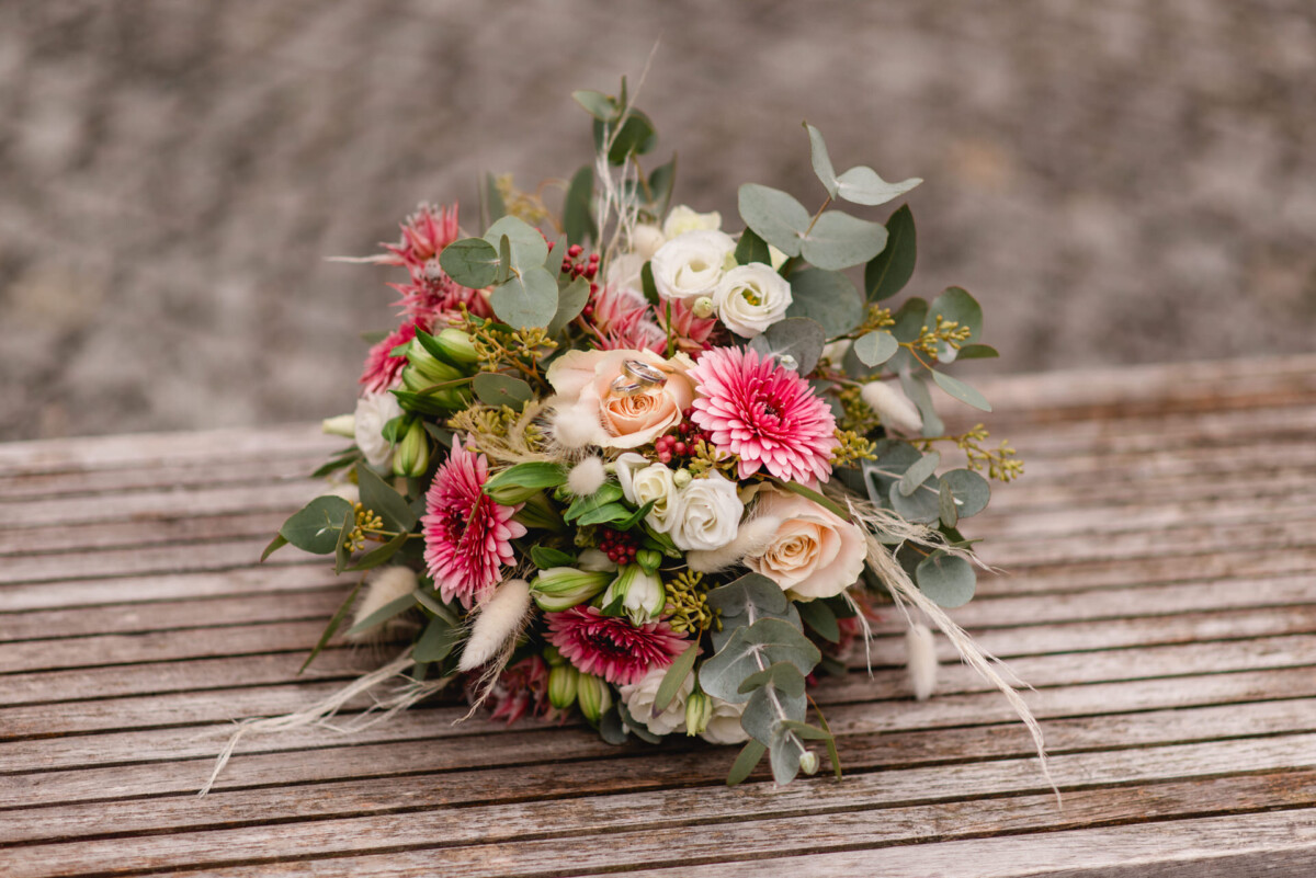 Ein Strauß rosa, weißer und pfirsichfarbener Blumen mit Grün steht auf einer Holzbank.