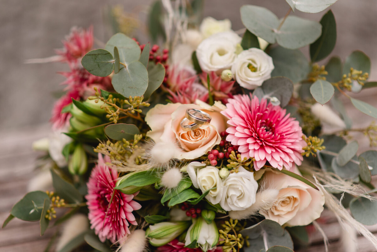 2024.10.05-Daniela-Daniel-Hochzeit-Gut-Schwabhof-Augsburg-00328 Ein lebendiger Strauß mit rosa Gerbera, pfirsichfarbenen Rosen, weißen Blüten, grünen Blättern und zwei silbernen Ringen zwischen den Blüten.