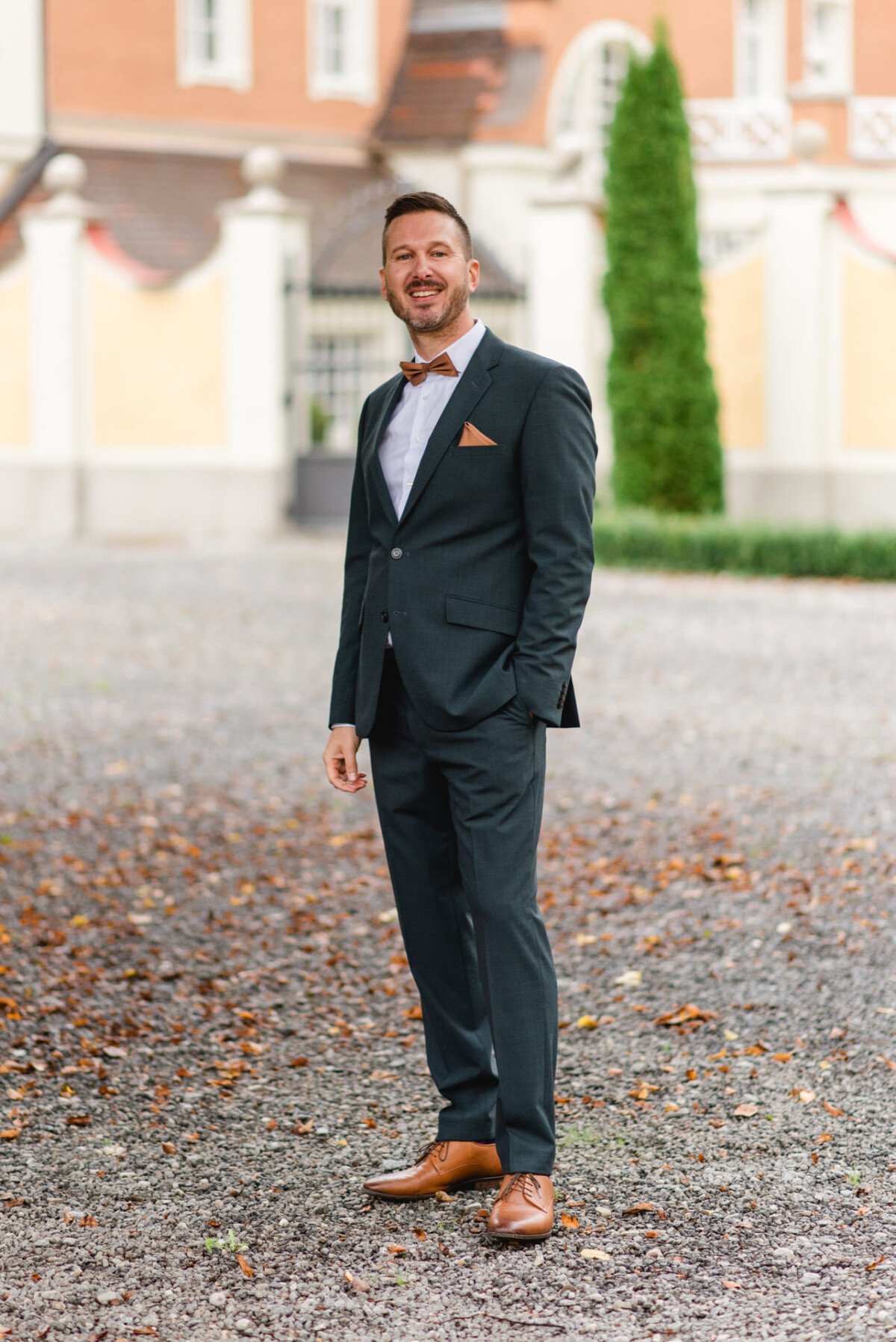 Ein Mann im Anzug steht draußen auf einem Kiesweg, umgeben von Herbstlaub, und lächelt in die Kamera.