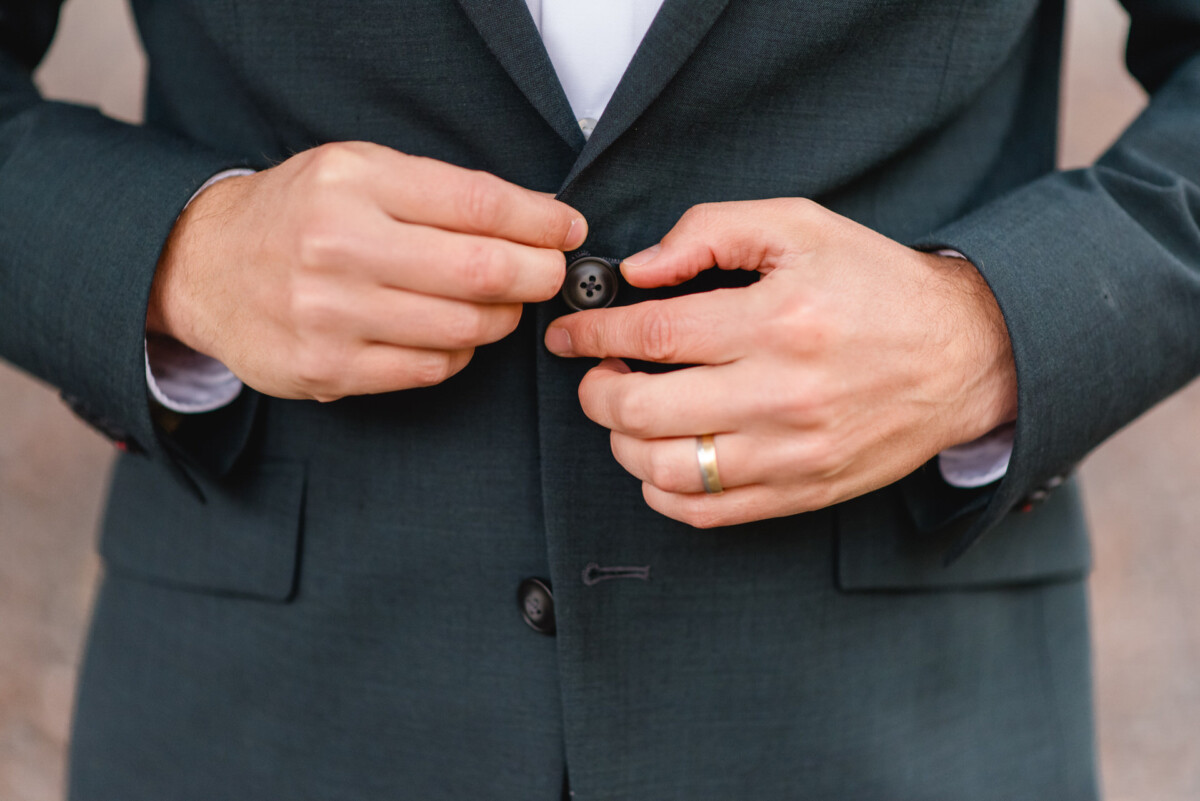 Eine Person im dunklen Anzug schließt einen Knopf an ihrem Jackett. An ihrer linken Hand trägt sie einen goldenen Ring.