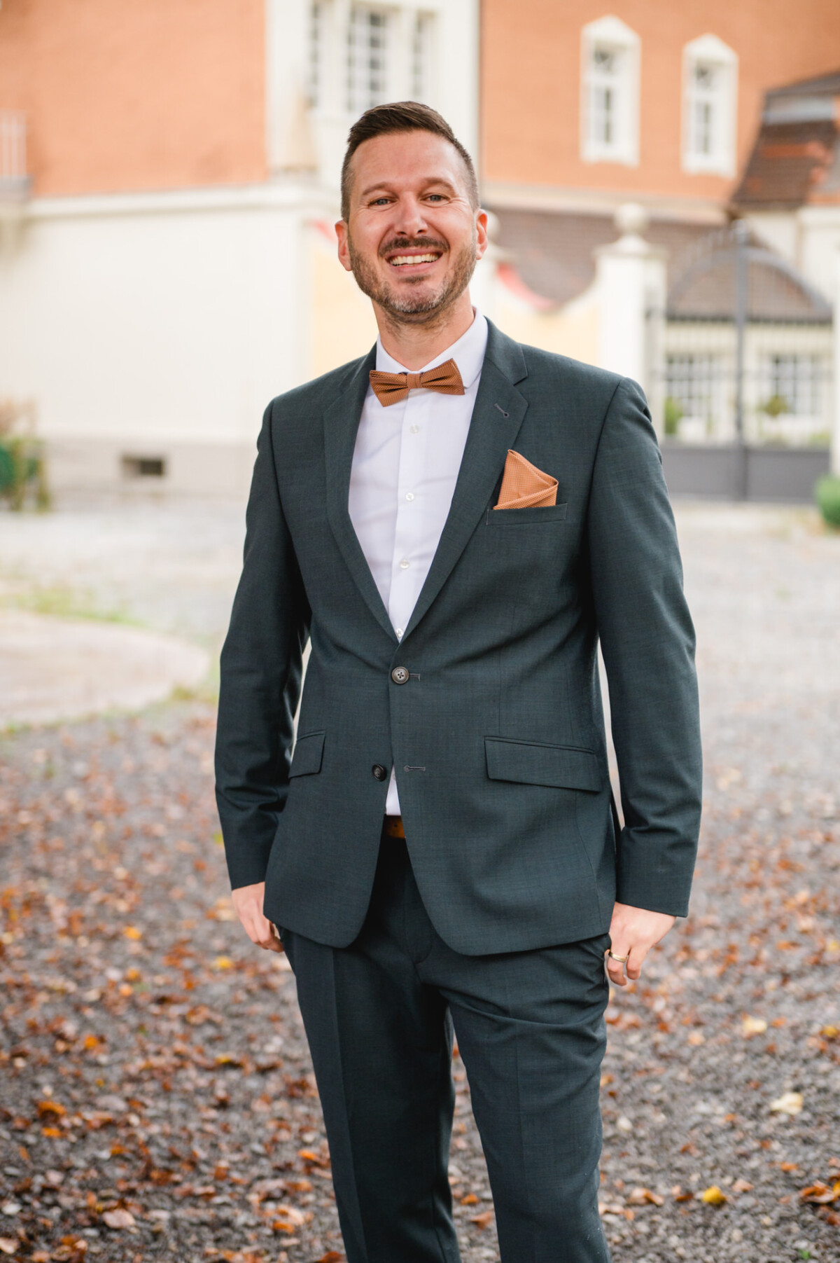 Ein Mann im dunklen Anzug mit hellbrauner Fliege und Einstecktuch steht auf einem Kiesweg vor einem Gebäude.