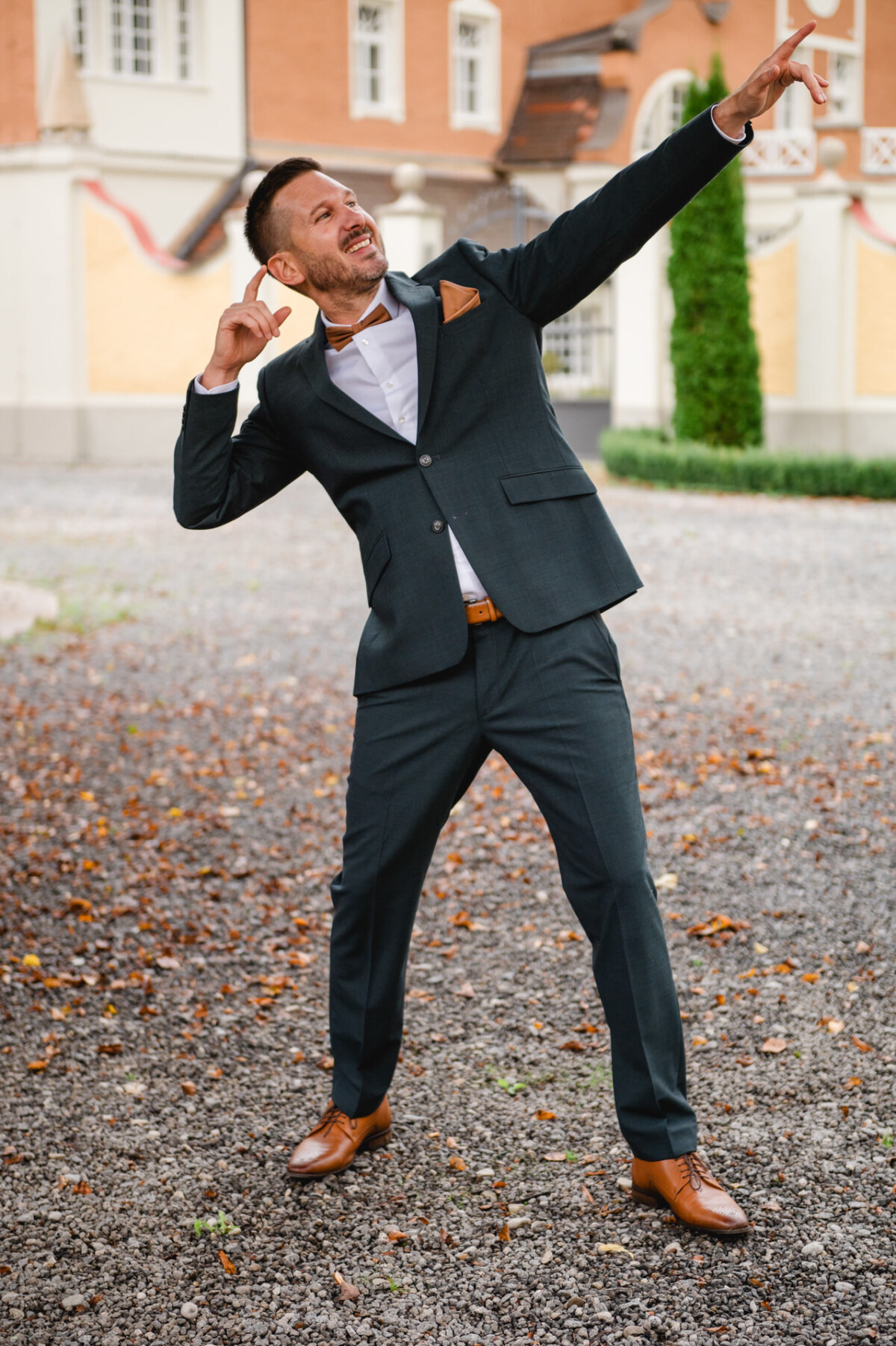 Ein Mann im dunklen Anzug macht eine feierliche Pose auf einem Kiesweg, im Hintergrund ein Gebäude und eine grüne Hecke.