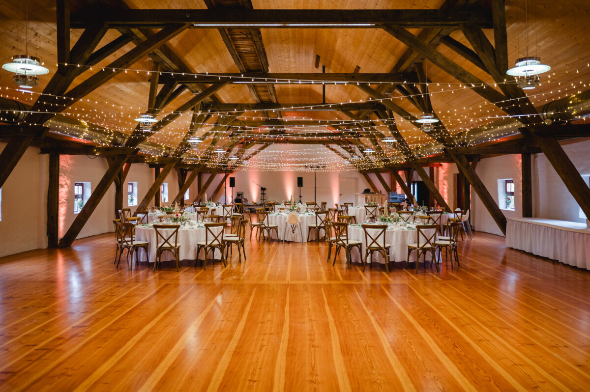 Geräumiger Veranstaltungssaal mit Holzbalken und Holzboden, dekoriert mit Lichterketten. Runde Tische und Stühle sind ordentlich mit Essbesteck angeordnet.