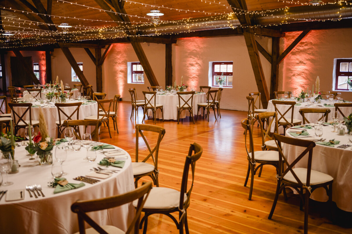 Ein warm beleuchteter Bankettsaal mit runden Tischen, die für eine Veranstaltung gedeckt sind. Die Tische sind mit weißen Tischdecken, Gläsern, Tellern und Blumendekorationen geschmückt. Über die Deckenbalken sind Lichterketten gespannt.