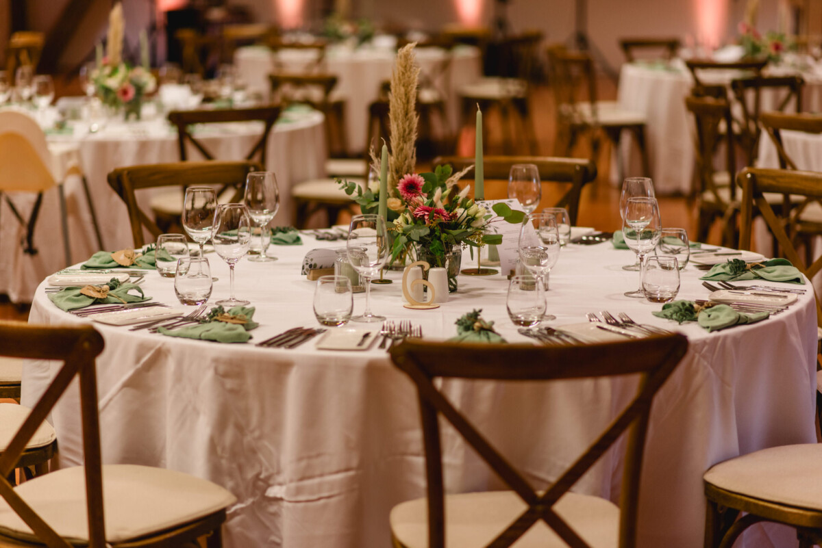 Runder Esstisch für eine formelle Veranstaltung, mit eleganten Blumendekorationen, hübsch arrangiertem Glasgeschirr und gefalteten Servietten auf einer weißen Tischdecke, mit Stühlen rund um den Tisch.