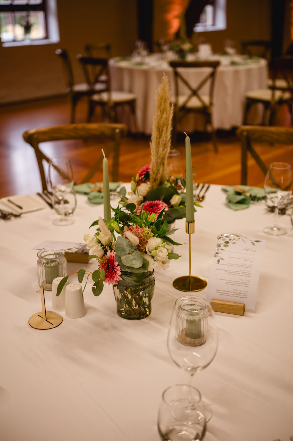 Ein dekorierter Tisch mit Blumenschmuck, grünen Kerzen, Weingläsern und einer Menükarte auf einer weißen Tischdecke.