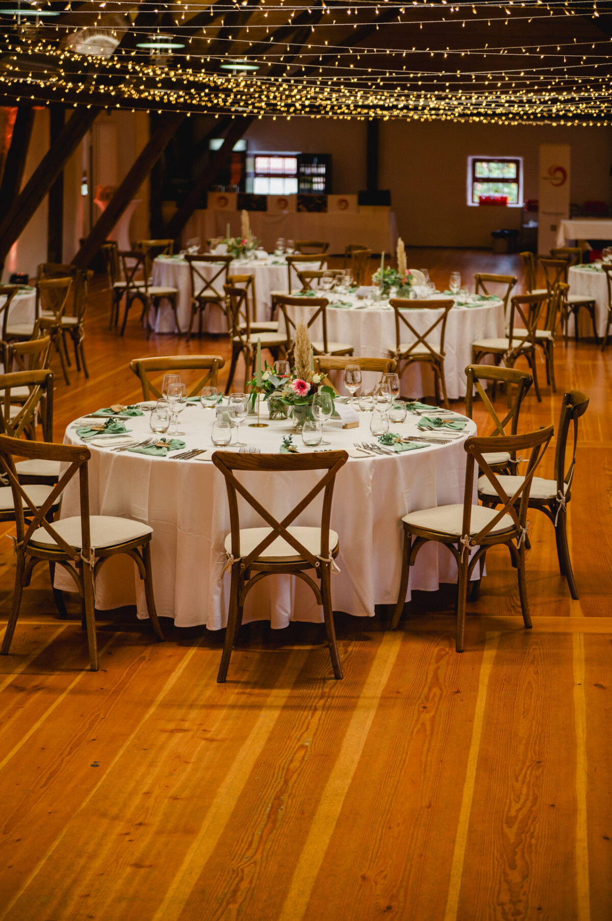 Ein dekorierter Bankettsaal mit runden Tischen mit weißen Tischdecken, Glaswaren und Blumendekorationen. Warme Lichterketten hängen von der Decke und schaffen eine elegante Atmosphäre.