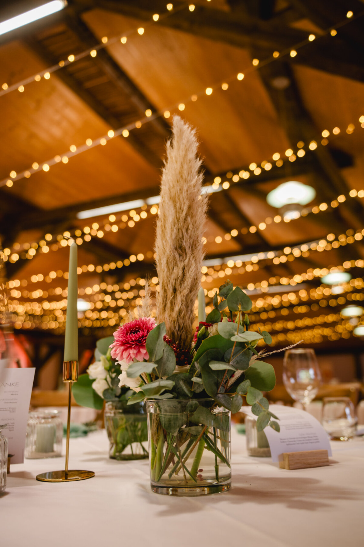 Ein floraler Tafelaufsatz mit Pampasgras, rosa Dahlien und Grünpflanzen in einer Glasvase, mit Kerzen und Lichterketten im Hintergrund.