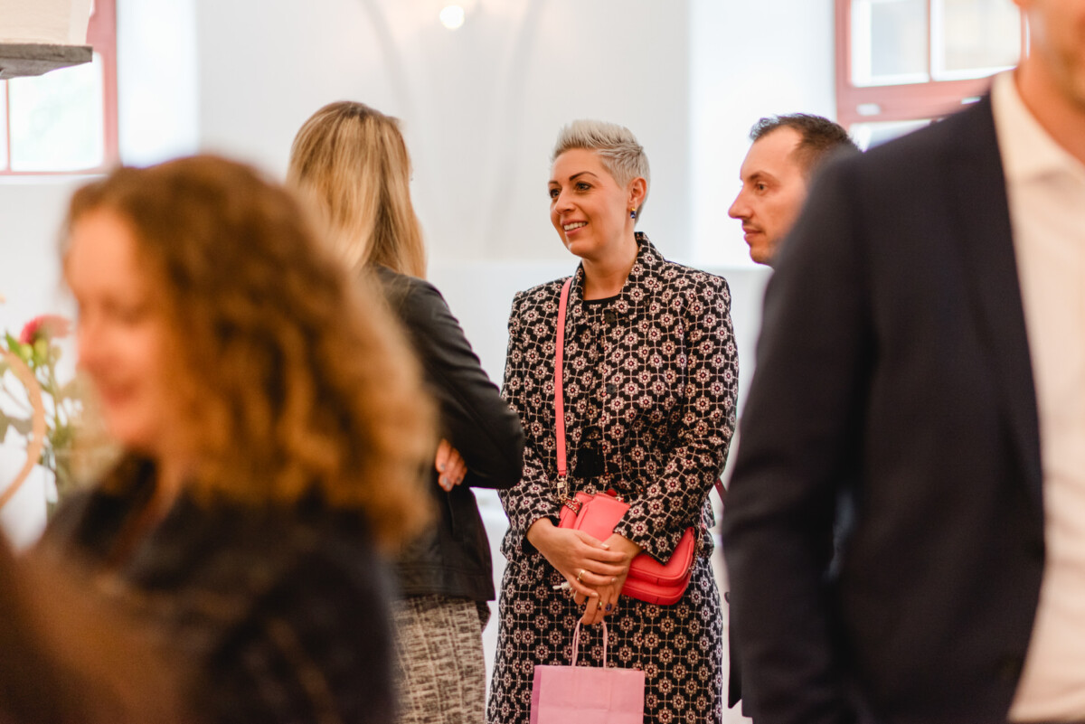 Eine Gruppe von Menschen unterhält sich im Innenbereich; eine Frau in einem gemusterten Kleid unterhält sich mit anderen und hält eine rosa Tasche.