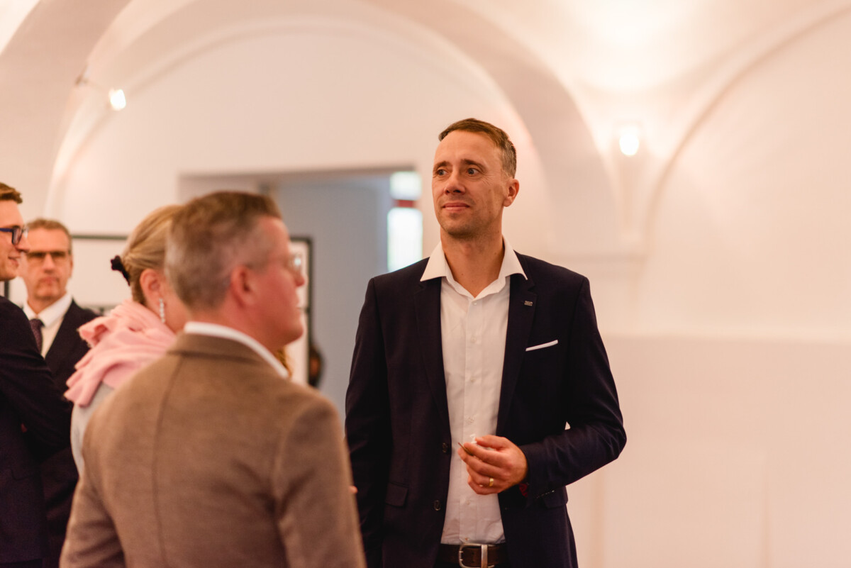 Ein Mann im Anzug steht in einem gut beleuchteten Raum mit Bögen, umgeben von anderen Menschen in Abendgarderobe.