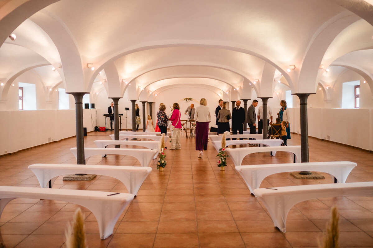 Eine Gruppe von Menschen steht und unterhält sich in einem geräumigen Saal mit gewölbter Decke, weiß bedeckten Bänken und Blumenarrangements.