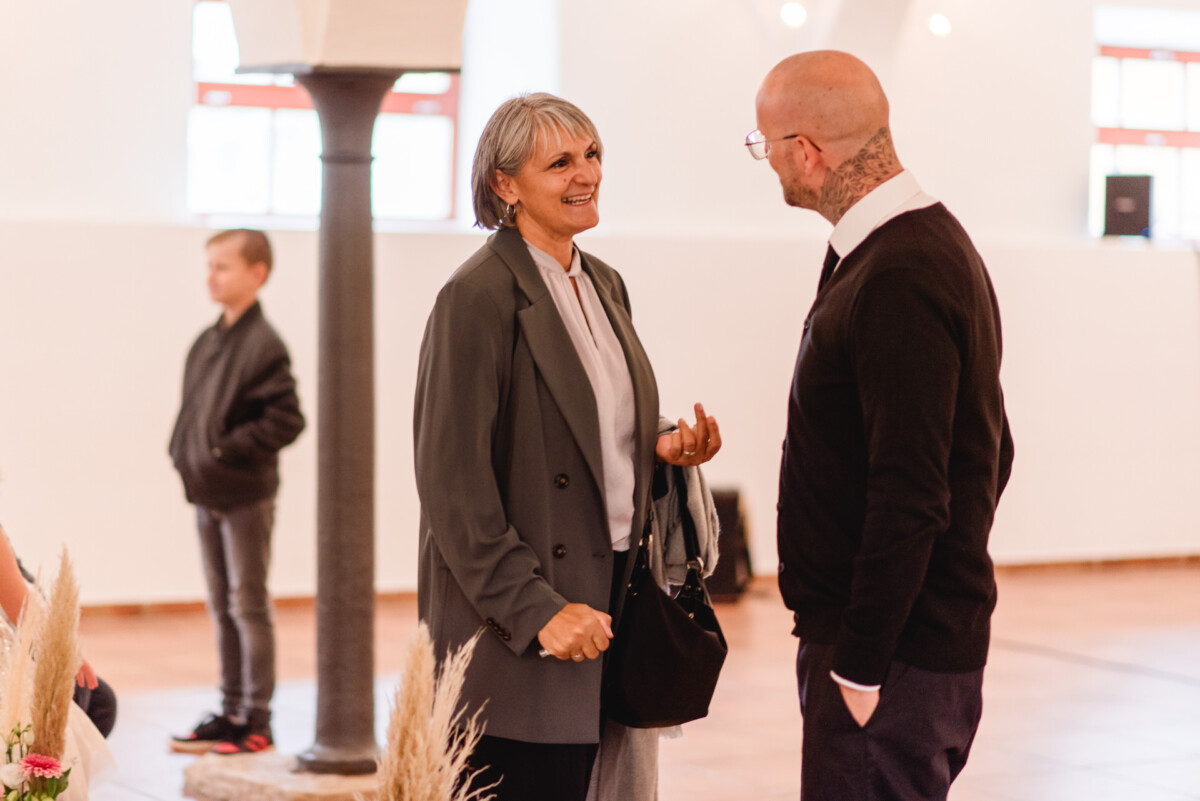 Zwei Personen unterhalten sich in einem Innenbereich. Im Hintergrund ist ein Kind zu sehen. Die Frau lächelt und hält eine Tasche.