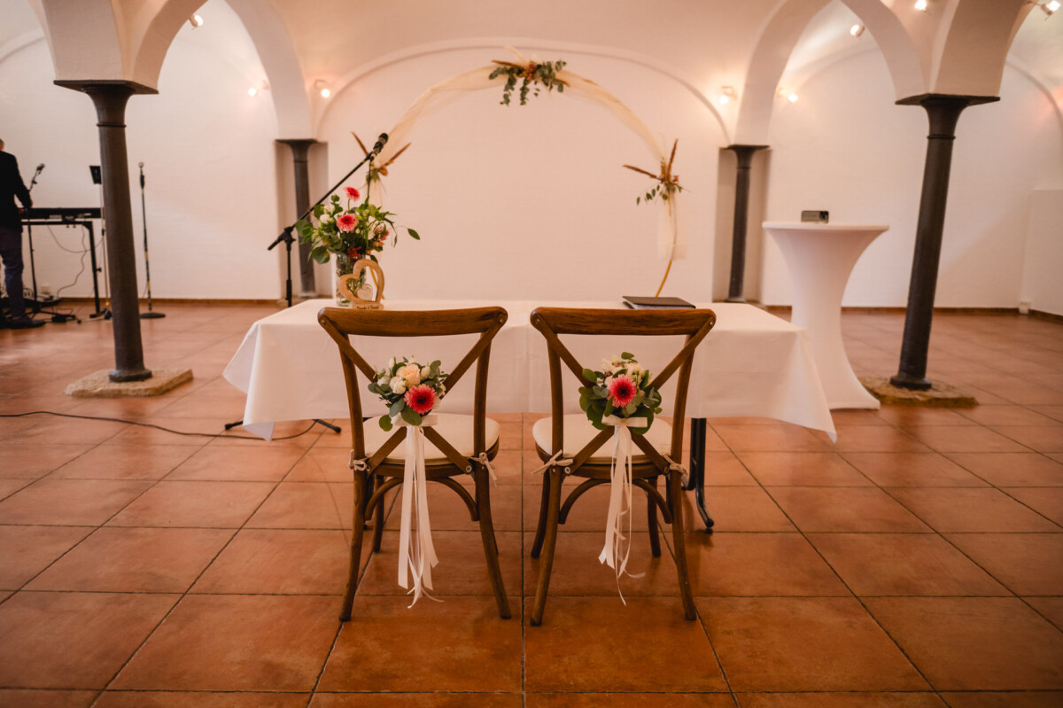 Aufbau einer Hochzeitszeremonie mit zwei mit Blumen und Bändern geschmückten Holzstühlen gegenüber einem Tisch und einem Blumenbogen in einem Raum mit gefliesten Böden und gewölbten Decken.