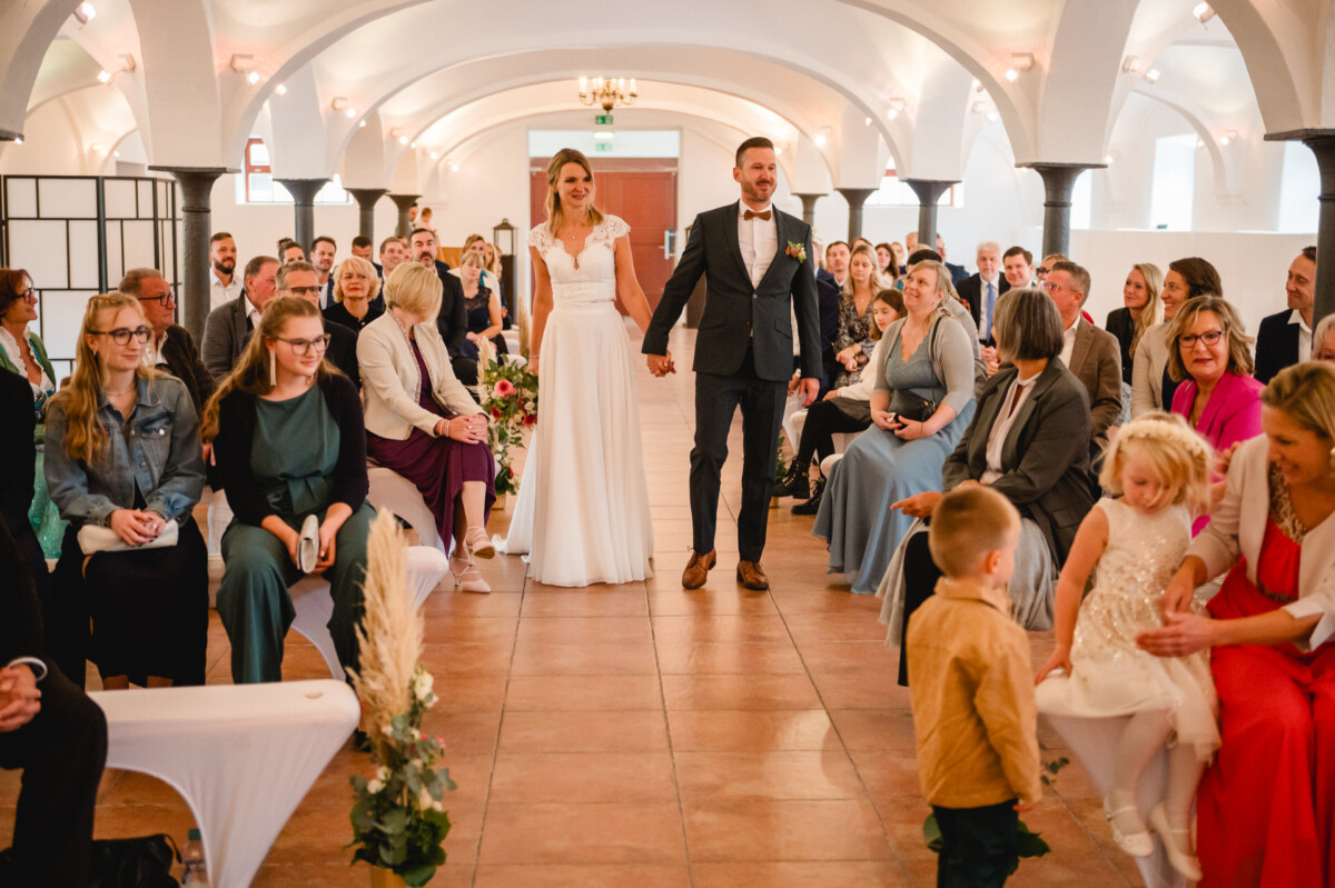 Braut und Bräutigam stehen in einem Raum voller sitzender Gäste. Ein Kind geht den Gang entlang. Die Kulisse ist durch gewölbte Decken und Dekor gekennzeichnet.