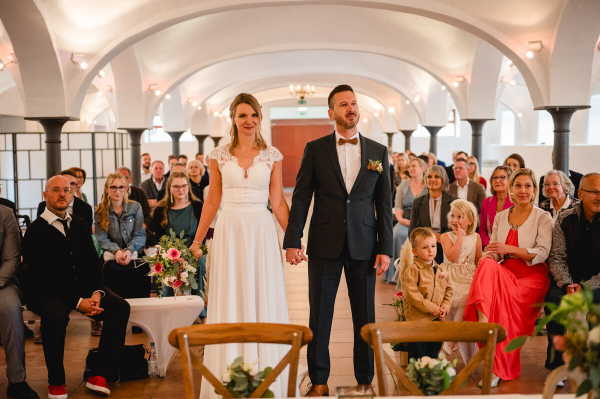 Braut und Bräutigam halten bei ihrer Hochzeitszeremonie in einem dekorierten Saal Händchen, umgeben von sitzenden Gästen, die ihnen zuschauen.