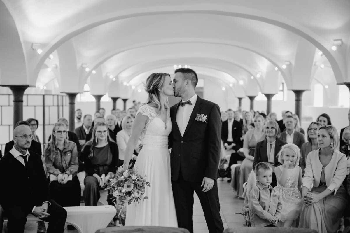 Braut und Bräutigam küssen sich bei ihrer Hochzeitszeremonie in einem Raum mit gewölbten Decken, umgeben von sitzenden Gästen. Ein Kind sitzt in der Nähe auf dem Boden.