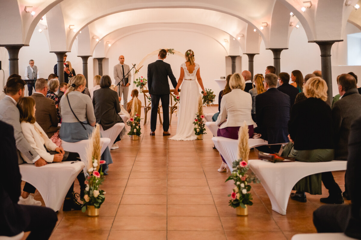Ein Paar steht während einer Hochzeitszeremonie in einem dekorierten Saal am Altar, auf beiden Seiten sitzen Gäste.