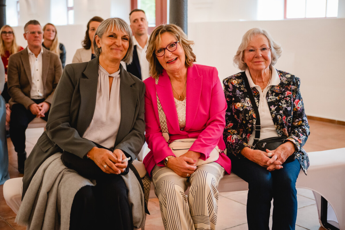 Drei Frauen sitzen lächelnd zusammen, andere sitzen im Hintergrund in einem hellen Raum.