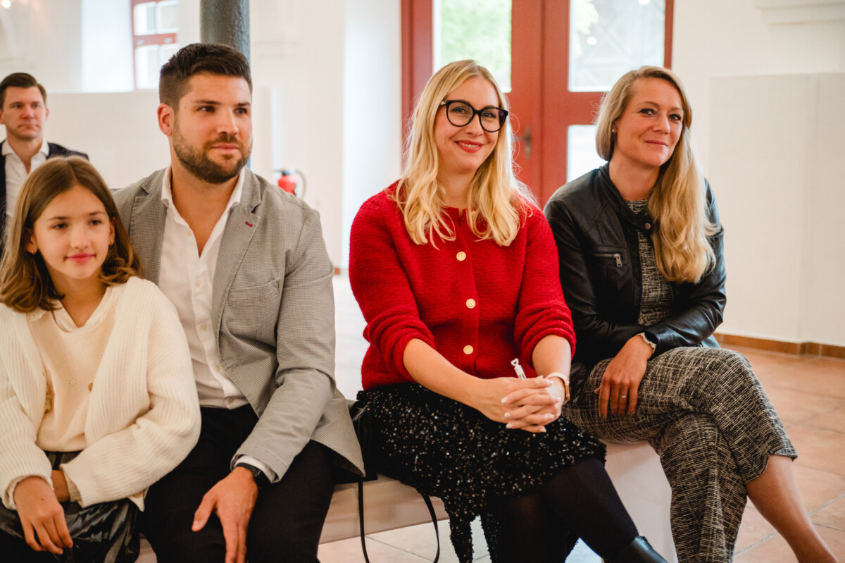 Eine Gruppe von vier Personen, darunter ein Kind, sitzt im Innenbereich mit einer roten Tür und großen Fenstern im Hintergrund.