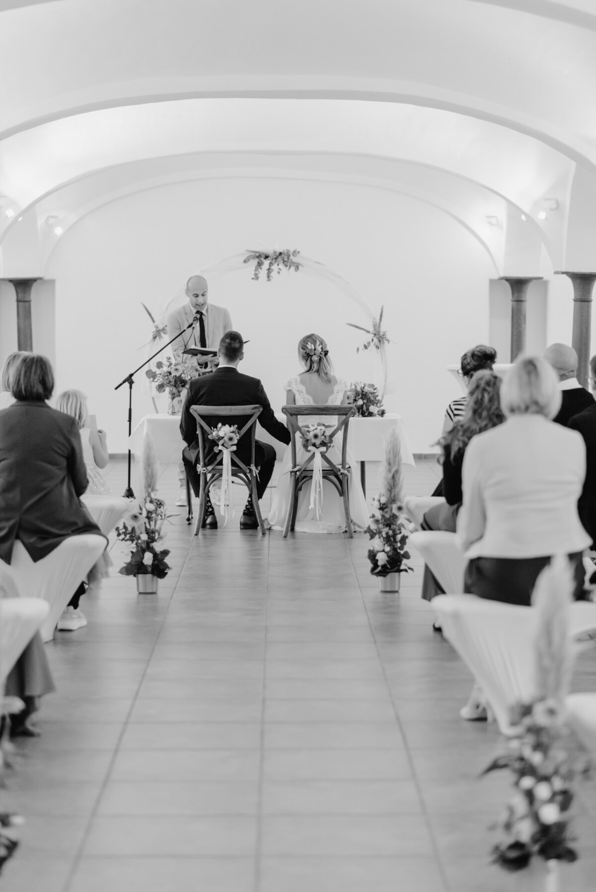 Ein Schwarzweißbild einer Hochzeitszeremonie in einer Kapelle. Das Paar sitzt dem Geistlichen gegenüber, die Gäste sitzen in Reihen. Blumenschmuck säumt den Mittelgang.