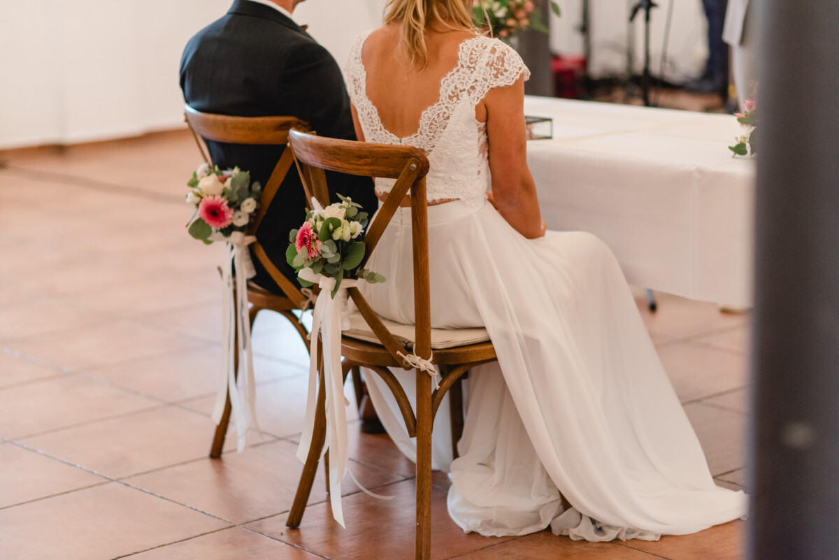 Ein Paar sitzt auf dekorierten Holzstühlen vor einem Tisch, vermutlich während einer Hochzeitszeremonie. Die Frau trägt ein weißes Spitzenkleid und an den Stühlen sind Blumenarrangements befestigt.