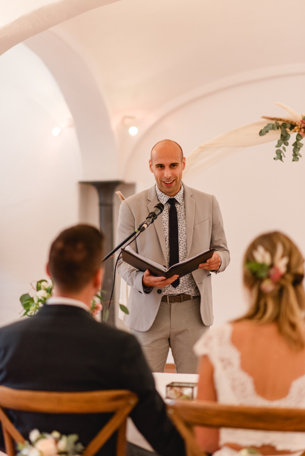 Ein Mann in einem hellgrauen Anzug spricht während einer Zeremonie in ein Mikrofon und steht einem sitzenden Paar gegenüber. Die Umgebung ist elegant mit Blumen geschmückt.