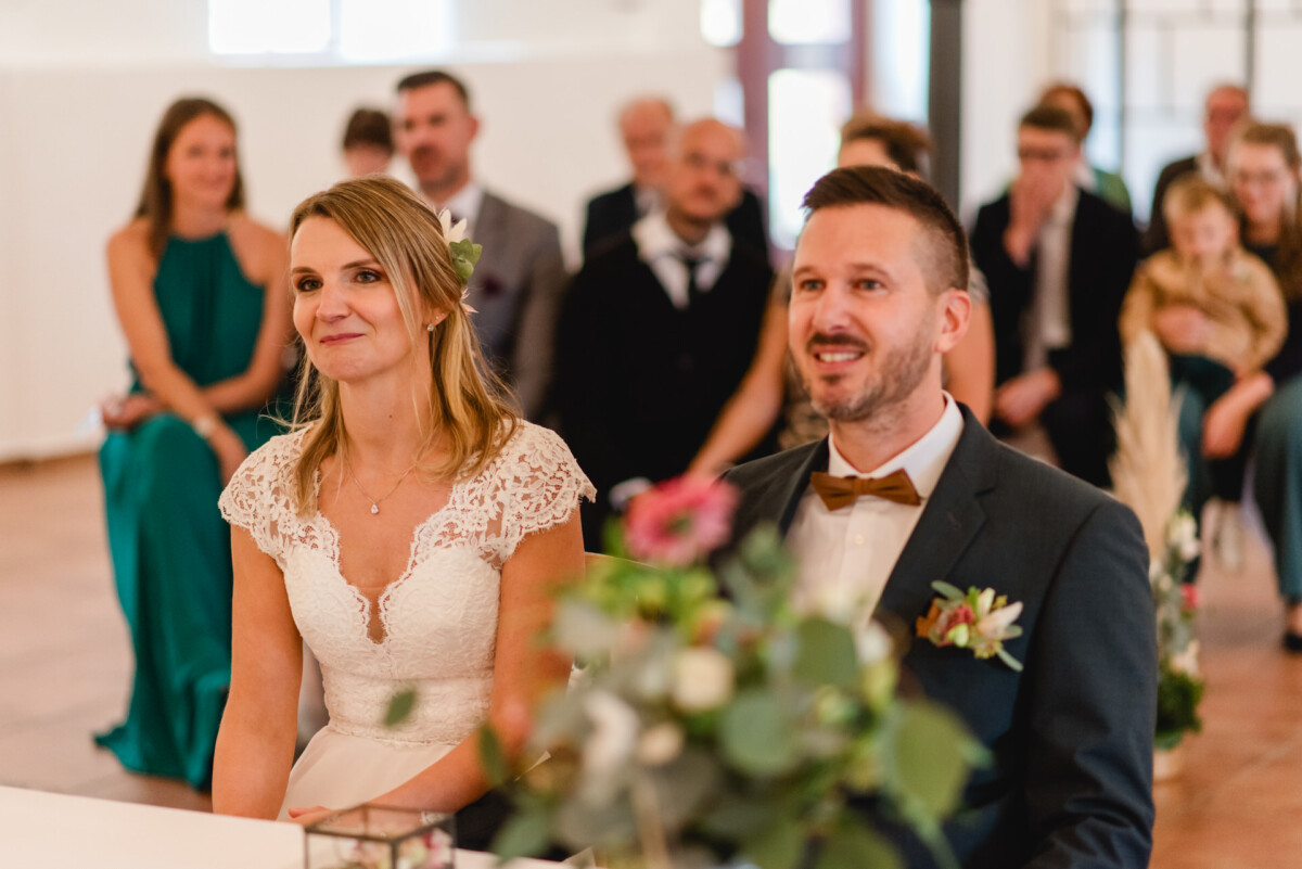 Braut und Bräutigam sitzen bei einer Hochzeitszeremonie zusammen. Vor ihnen steht ein dekorierter Tisch, im Hintergrund sitzen die Gäste.