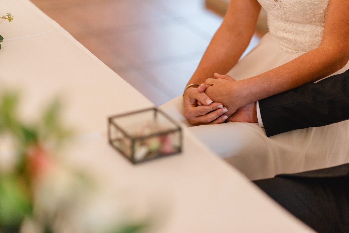 Braut und Bräutigam sitzen an einem Tisch und halten sich an den Händen. Im Vordergrund sind eine kleine dekorative Schachtel und ein Blumenarrangement zu sehen.