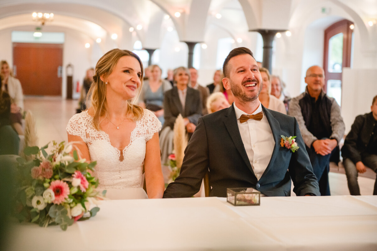 Ein Paar im Hochzeitskleid sitzt lächelnd bei einer Zeremonie in einem hellen Saal. Hinter ihnen sitzen Gäste.