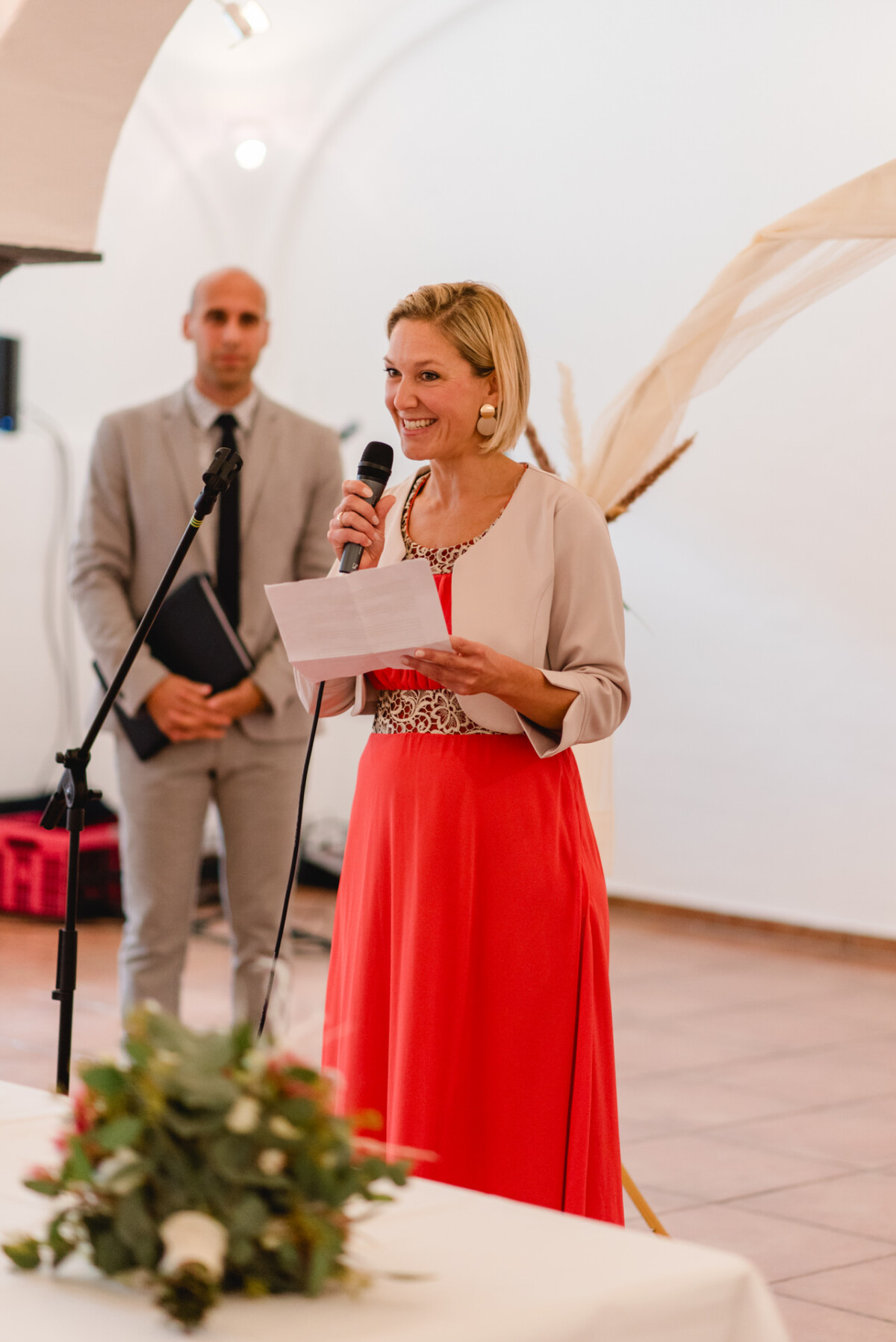 Eine Frau in einem roten Kleid spricht in ein Mikrofon und hält dabei ein Blatt Papier in der Hand. Im Hintergrund steht ein Mann in einem grauen Anzug. Im Vordergrund ist ein Blumenarrangement zu sehen.