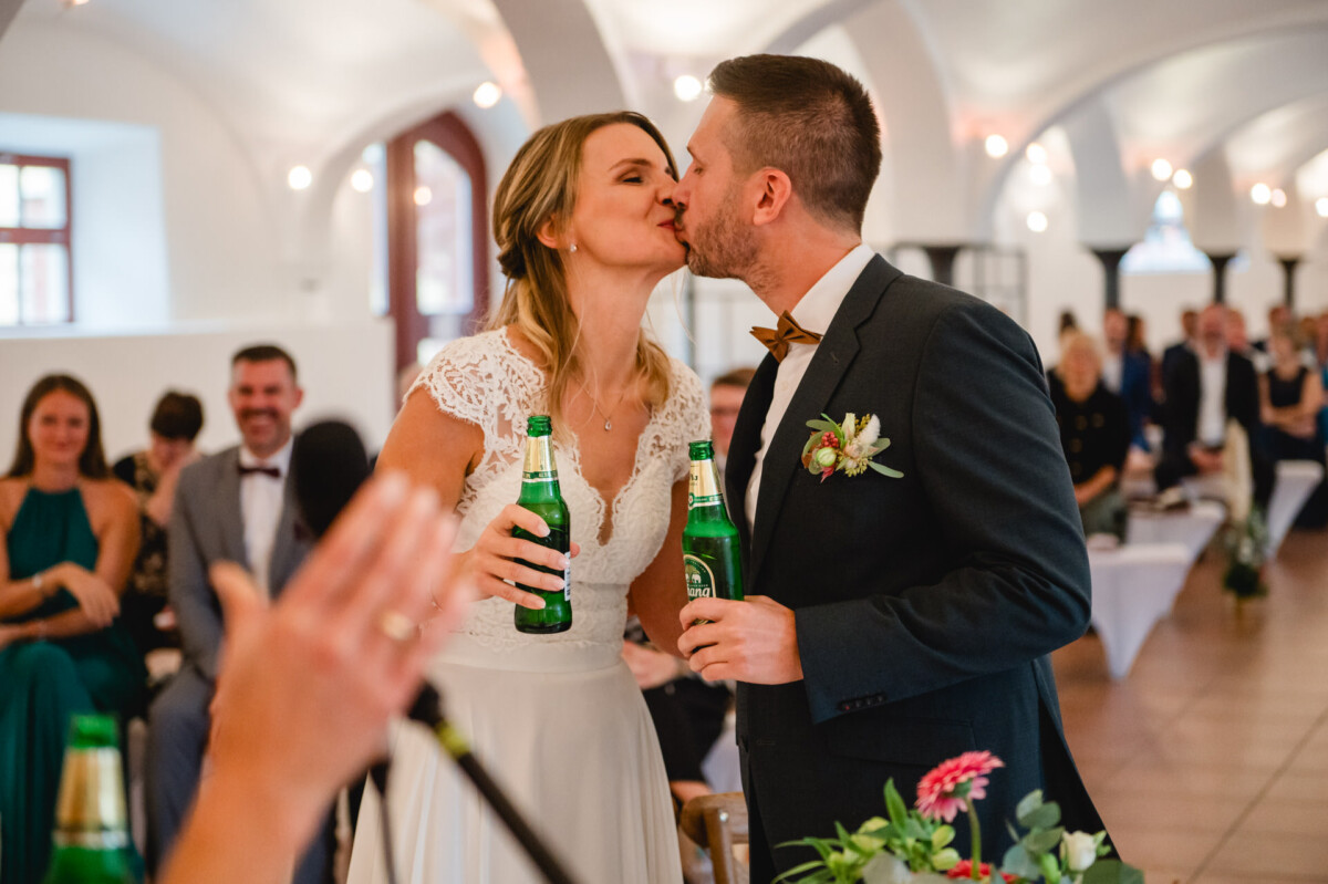 Bei einer Hochzeitszeremonie küssen sich Braut und Bräutigam, während sie Bierflaschen halten. Im Hintergrund sitzen die Gäste.