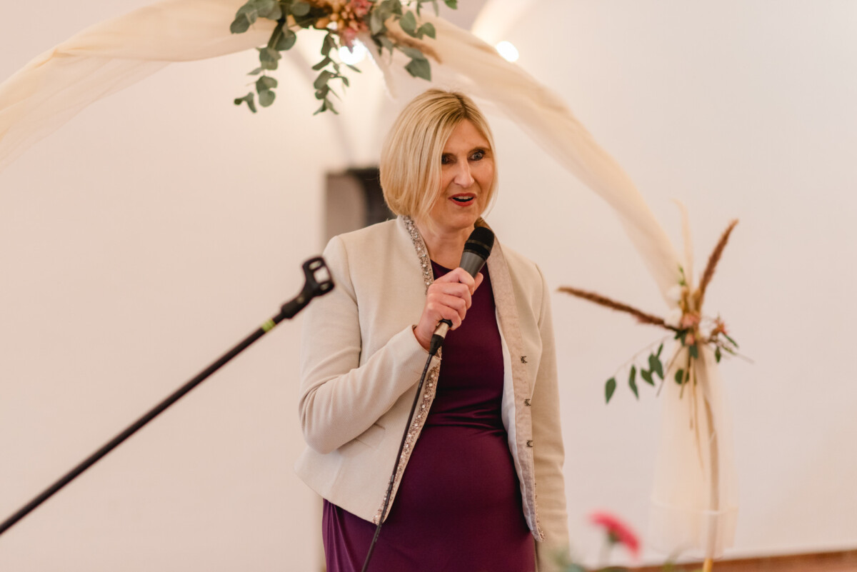 Eine Frau im Blazer spricht unter einem dekorierten Torbogen in ein Mikrofon.