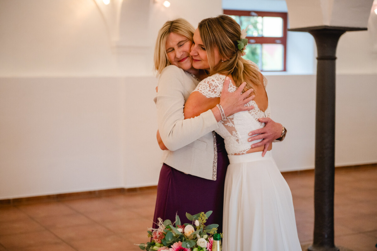 Zwei Frauen umarmen sich herzlich im Innenbereich. Eine trägt ein weißes Kleid und hält einen Blumenstrauß in der Hand.