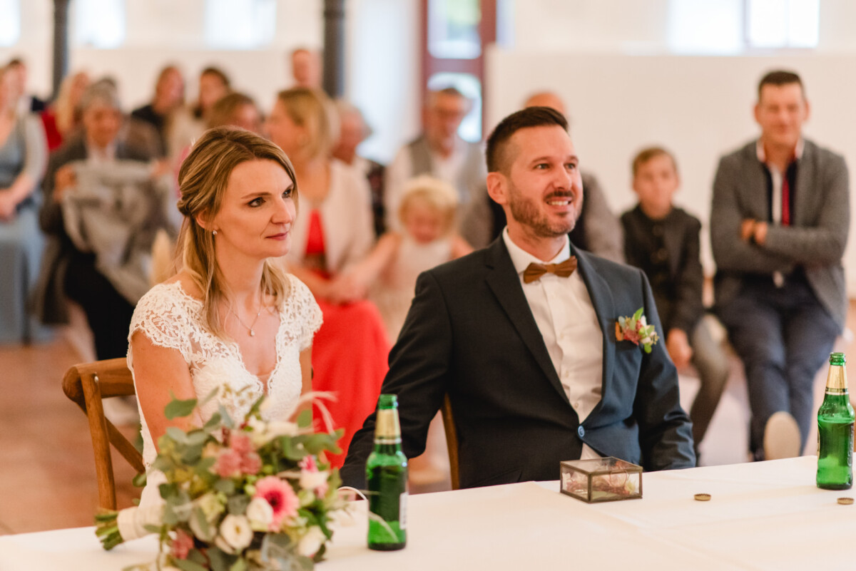 Ein Paar sitzt in Abendgarderobe an einem Tisch, vor ihnen ein Blumenstrauß und Flaschen. Im Hintergrund sitzt aufmerksam das Publikum.