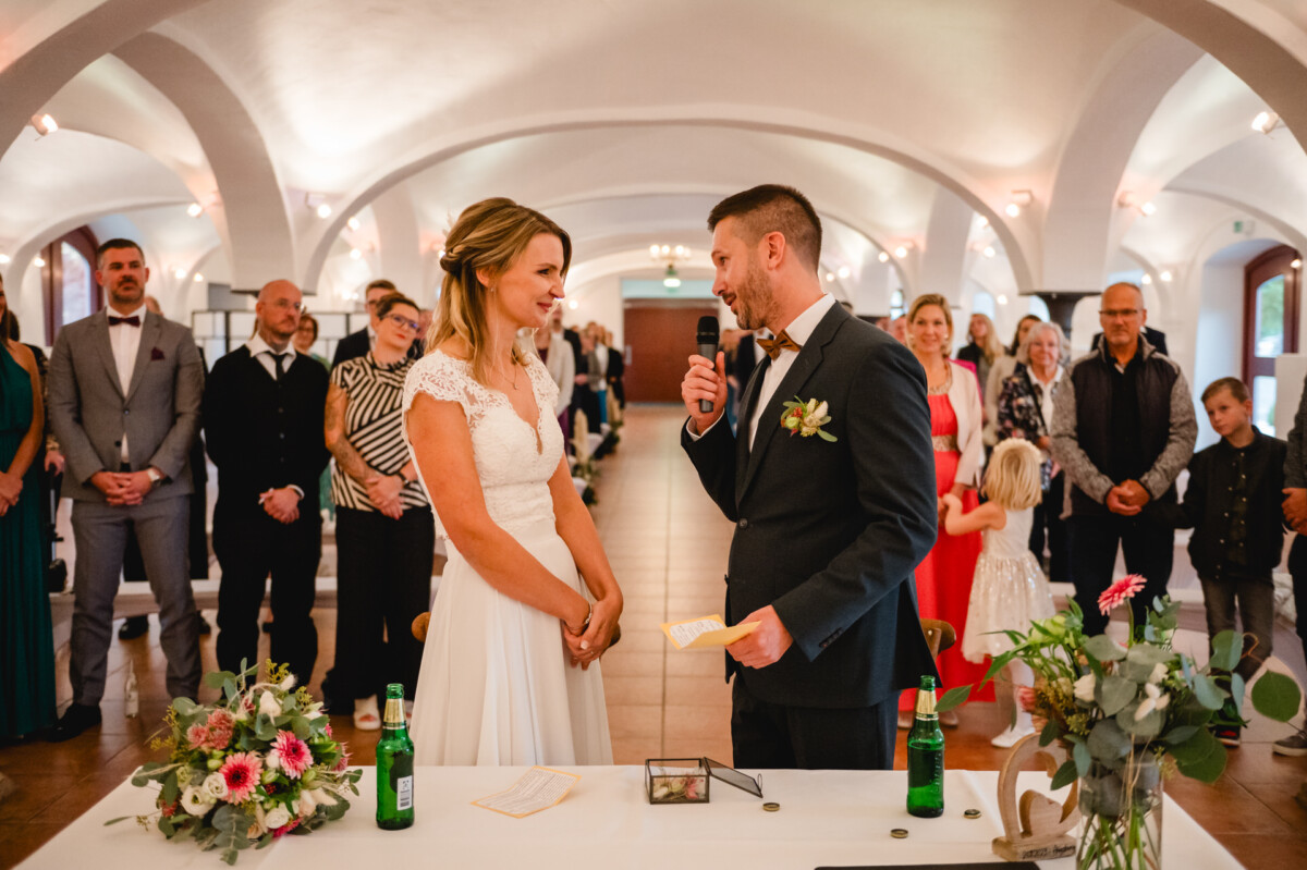 Braut und Bräutigam stehen während einer Hochzeitszeremonie an einem Tisch und lächeln einander an. Im Hintergrund sitzen und stehen Gäste, auf dem Tisch stehen Blumen und Flaschen.