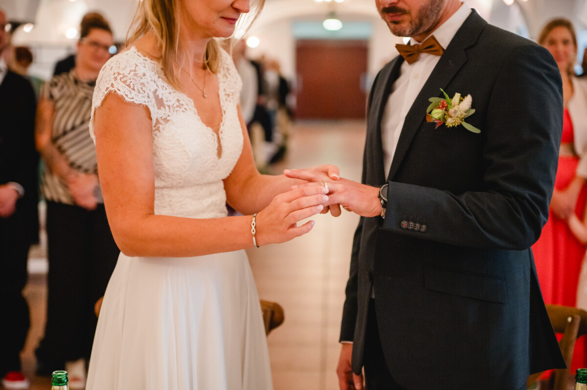 Während einer Hochzeitszeremonie steckt eine Braut dem Bräutigam einen Ring an den Finger. Sie stehen im Haus, umgeben von Gästen.