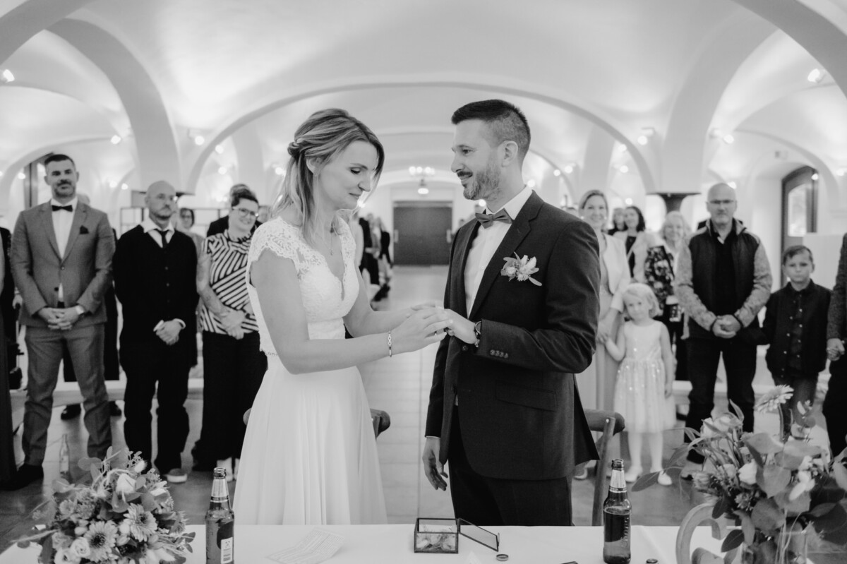 Während einer Hochzeitszeremonie steckt eine Braut dem Bräutigam einen Ring an den Finger, umgeben von Gästen in einem elegant dekorierten Raum.