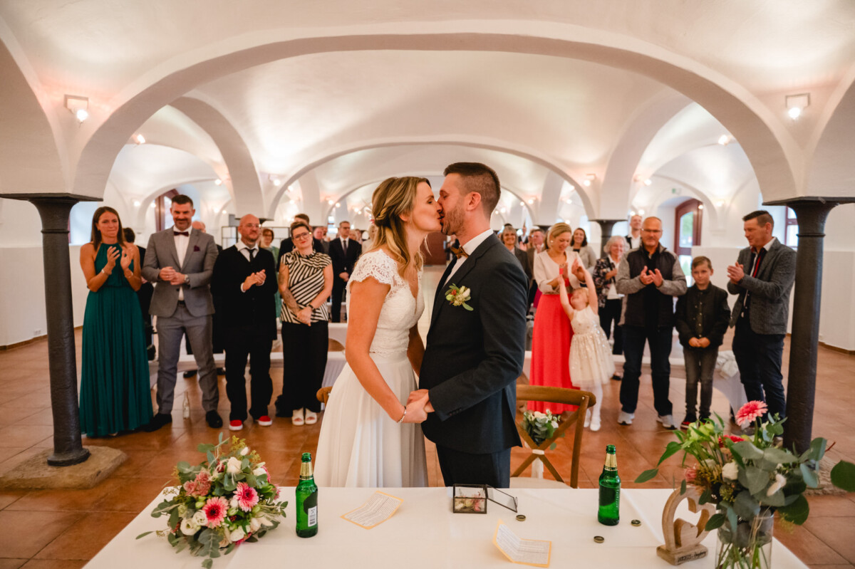 Braut und Bräutigam küssen sich in einem Raum, während die Gäste klatschen und zuschauen. Im Vordergrund steht ein Tisch mit Blumen und Flaschen.