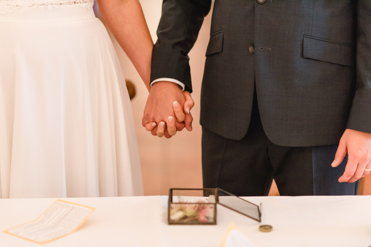 Ein Paar hält sich in einem formellen Rahmen an den Händen. Es trägt einen Anzug und ein weißes Kleid. Auf dem Tisch vor ihnen liegt eine kleine Glasschachtel mit Blumen und einem Ring.